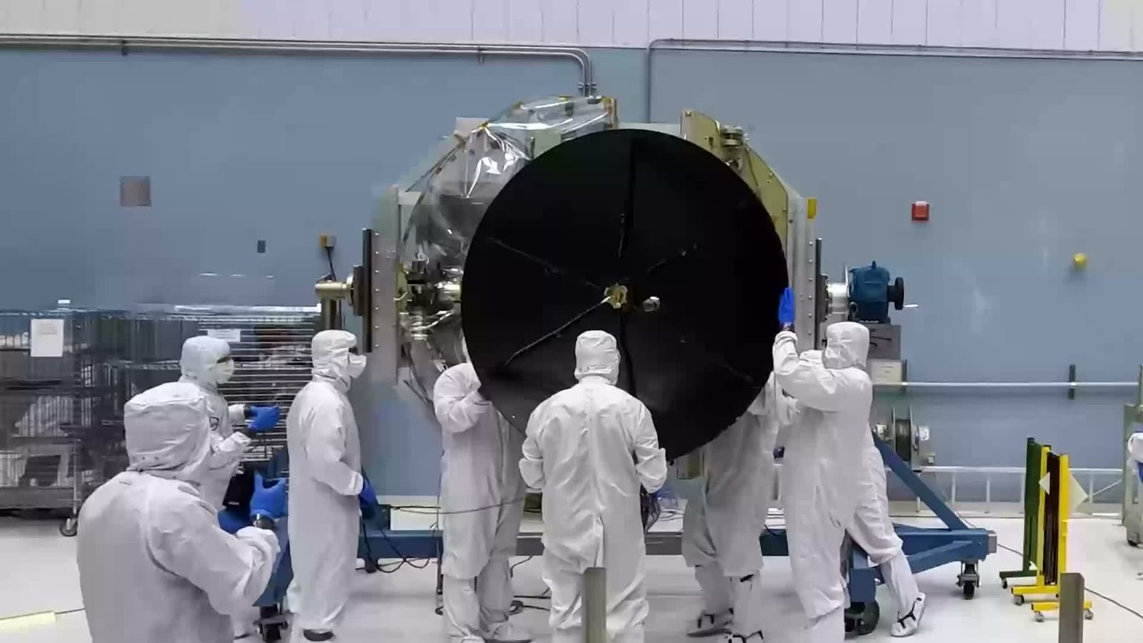Technicians in clean-room suits fitting a large dish antenna to a spacecraft